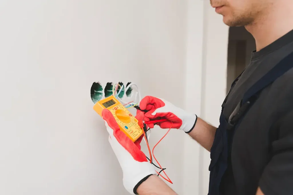 Electrician in black shirt and red gloves holding a yellow digital multimeter against white outlet box to test electrical voltage.