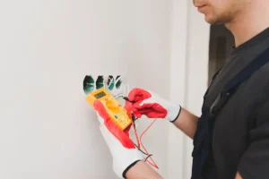 Electrician in black shirt and red gloves holding a yellow digital multimeter against white outlet box to test electrical voltage.