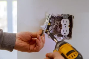 Electrician installing a light switch into a wall outlet box using a power drill and screwdriver.