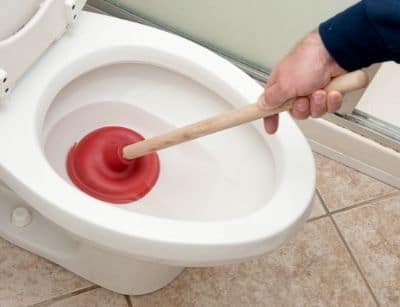 Person using a red plunger to unclog a white toilet bowl in a bathroom with tile flooring.