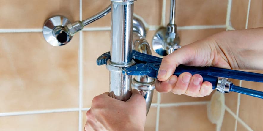 Plumber's hands using a blue wrench to tighten a chrome pipe connection under a bathroom sink against tiled wall.