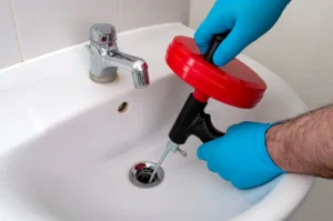 Person wearing blue gloves uses a red and black drain snake tool to clear a clogged bathroom sink drain.