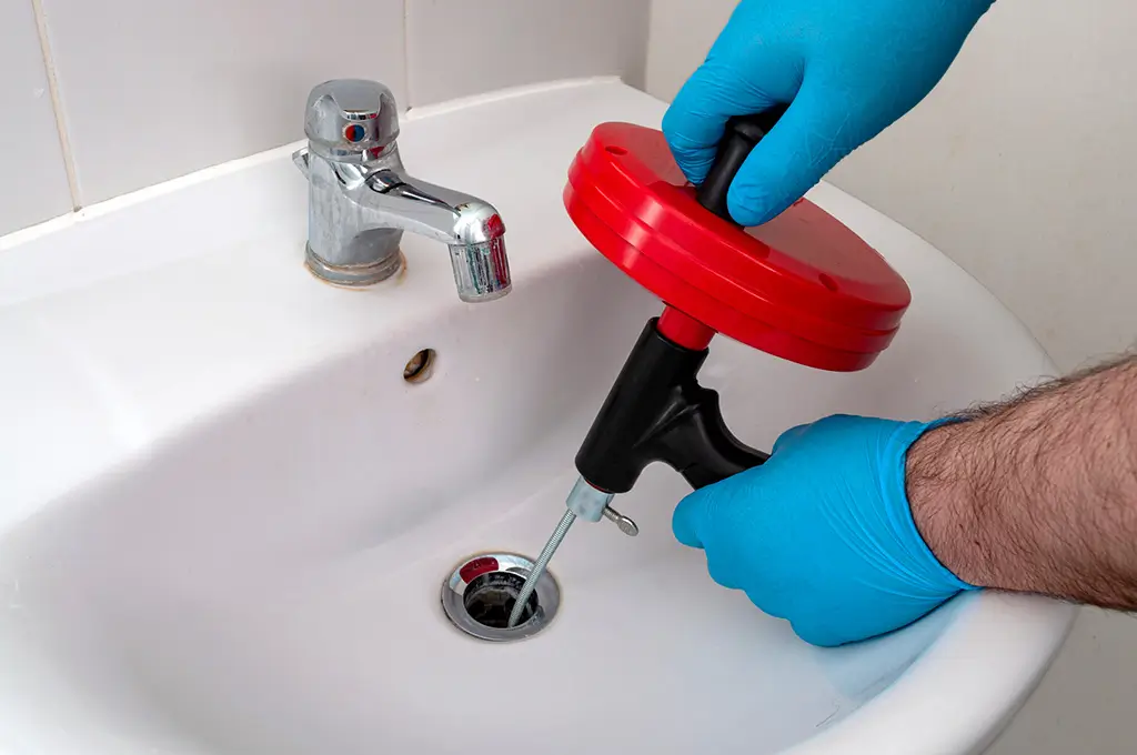 Person wearing blue gloves uses a red and black drain snake tool to clear a clogged bathroom sink drain.