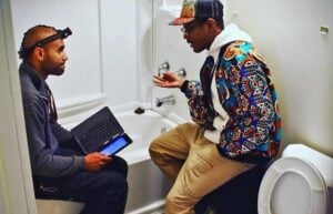 Two men having a discussion in a bathroom next to a toilet, one holding a tablet device while the other gestures during conversation.