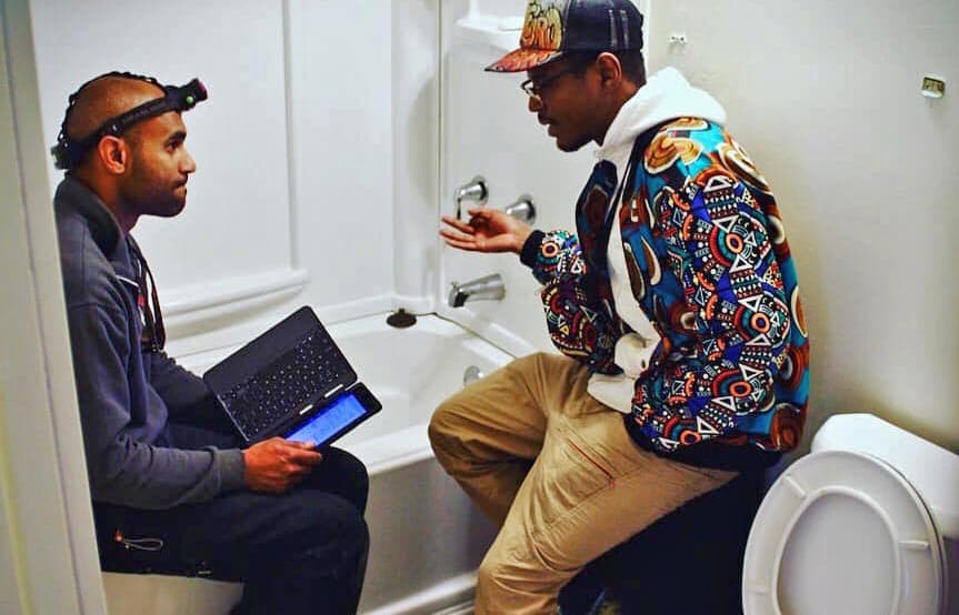 Two men having a discussion in a bathroom next to a toilet, one holding a tablet device while the other gestures during conversation.