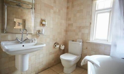 Bright bathroom featuring a pedestal sink, toilet, bathtub, and shower enclosure with beige tile walls and natural light from a window.