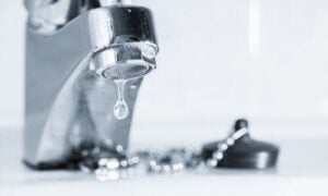 A chrome faucet drips water with a single droplet suspended below the aerator against a blurred background.