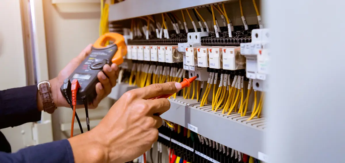 Electrician holding clamp meter while testing white circuit breakers in electrical panel with yellow wires.