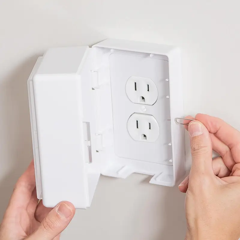Hands installing a white childproof safety cover over electrical outlets on a wall.