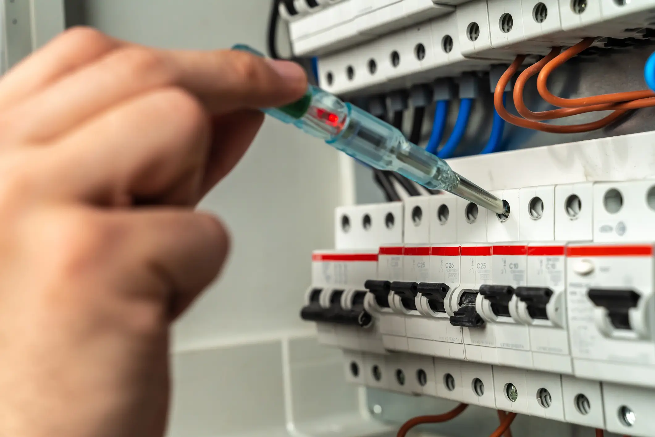 Electrician holding a voltage tester probe near circuit breakers labeled C25 on an electrical panel with colored wiring.