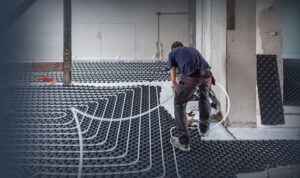 Worker installing radiant heating tubes on black insulation mat in an unfinished room with white walls and support columns.