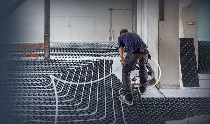 Worker installing radiant heating tubes on black insulation mat in an unfinished room with white walls and support columns.