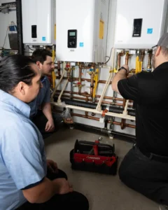 Two technicians inspect copper piping and controls on installed tankless water heaters mounted on wall.