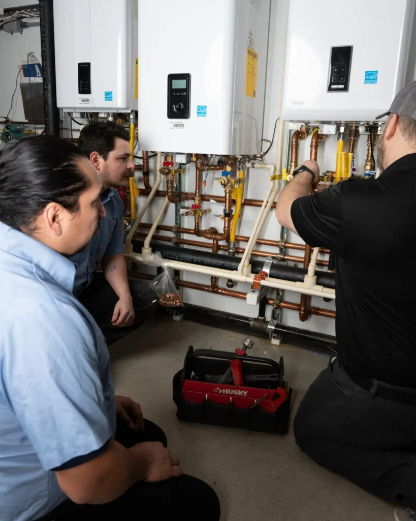 Two technicians inspect copper piping and controls on installed tankless water heaters mounted on wall.