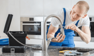 Professional plumber in blue overalls repairs a modern kitchen faucet over a countertop with tools and supplies nearby.