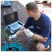 Technician examines water pipes and equipment during leak detection and repair inspection on gravel ground.