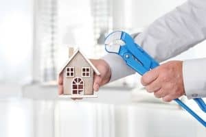 Hands holding blue wrench tools working on miniature wooden house model indoors.