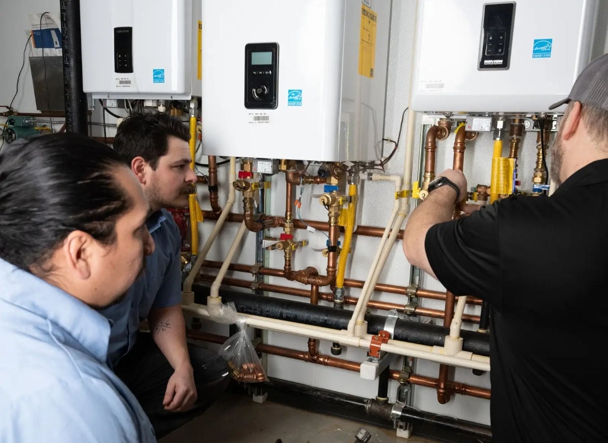 Two technicians examining tankless water heater piping system with copper and PVC connections during installation or maintenance work.