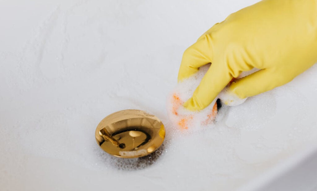 Gloved hand scrubbing a gold drain cover with a cleaning brush and soapy water in a white bathtub.
