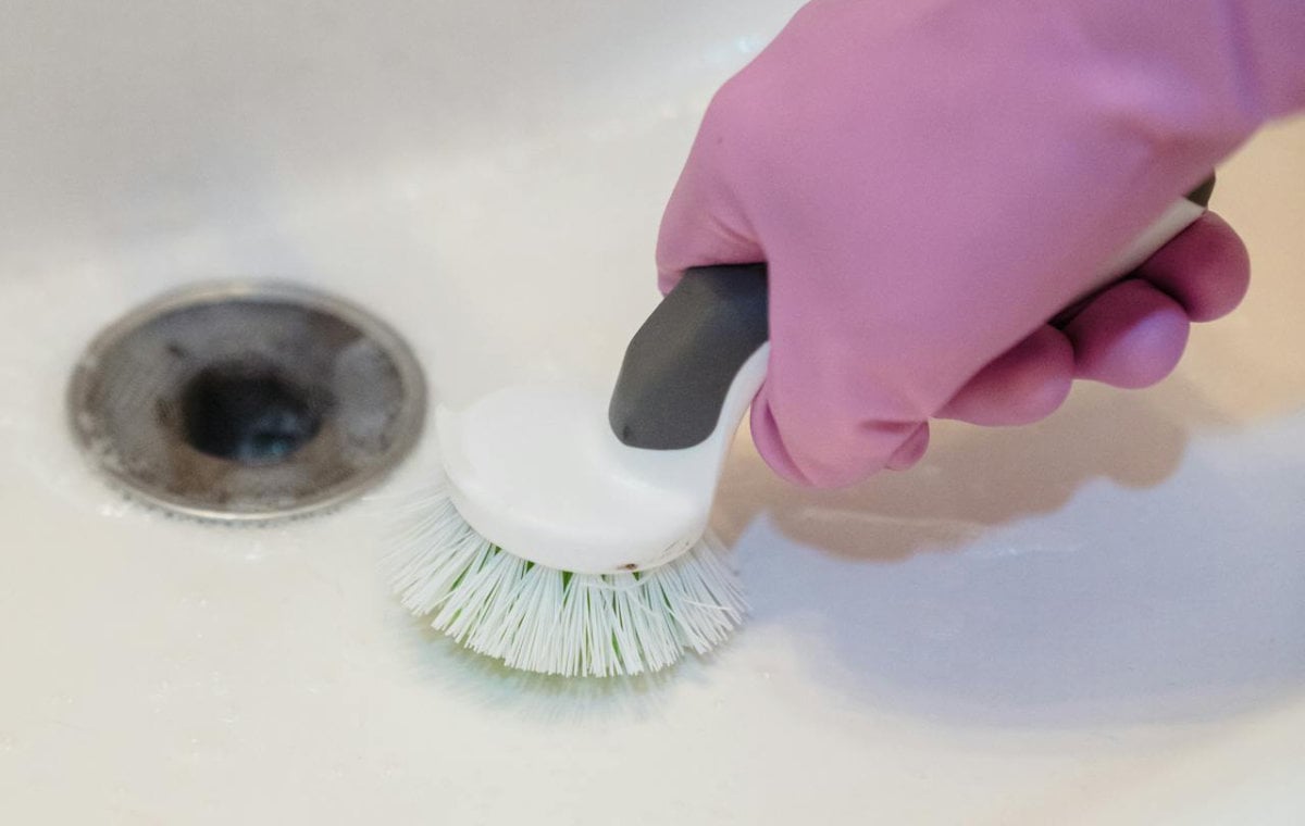 Person wearing pink gloves using a white-handled brush to clean a kitchen sink drain opening.
