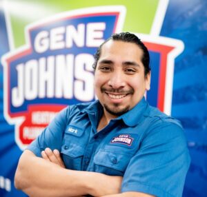 Smiling man in blue Gene Johns work uniform with arms crossed against branded backdrop.