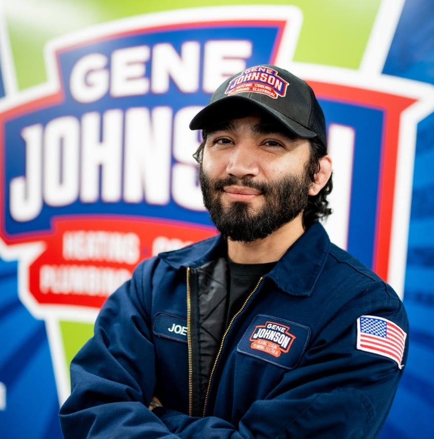 Man wearing navy Gene Johnson branded jacket and black cap with American flag patch, smiling at camera against blurred signage.