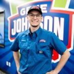 Young man wearing glasses and a blue work shirt with name tag smiling in front of branded signage.