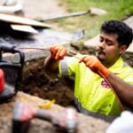 Male construction worker wearing neon yellow safety vest and orange gloves operates equipment in an excavation pit at a job site.