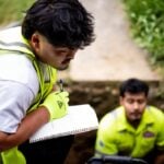 Two construction workers wearing high-visibility vests document inspection findings on a spiral notebook at an outdoor job site.
