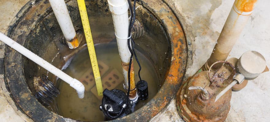 Black sump pump with discharge pipe and check valve installed inside a concrete basement pit with water and rust staining.