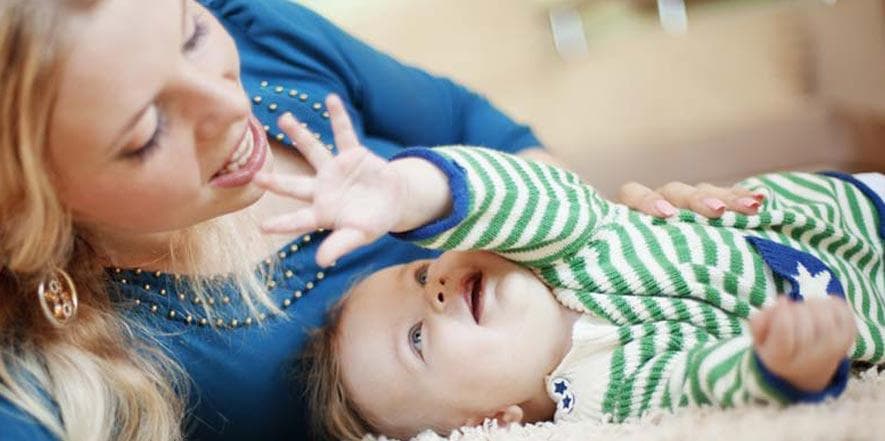 Woman playing with smiling baby wearing green striped clothing on carpet. Both appear happy and engaged in playtime together.