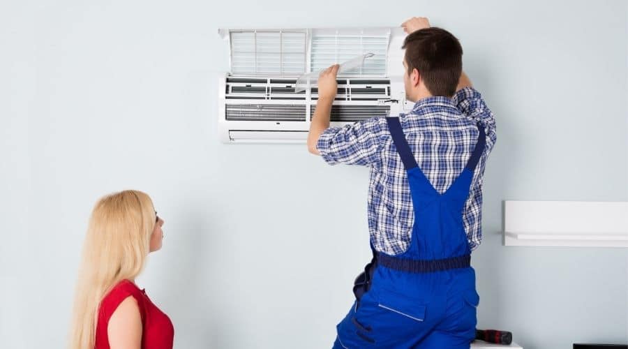 Technician in blue overalls inspecting a wall-mounted air conditioning unit while a woman observes.