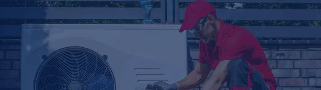 Technician in red uniform and cap installing or servicing an outdoor air conditioning unit with visible fan component.