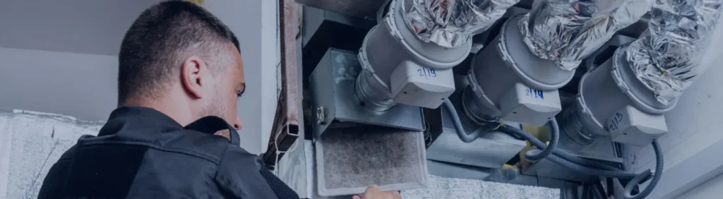 Technician in black jacket examining large cylindrical HVAC components with white labels and flexible ductwork connections.