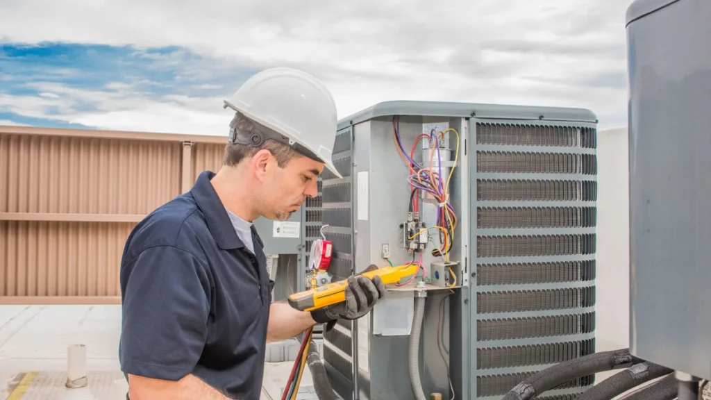 HVAC technician in hard hat and work gloves using a multimeter to diagnose an outdoor air conditioning unit.