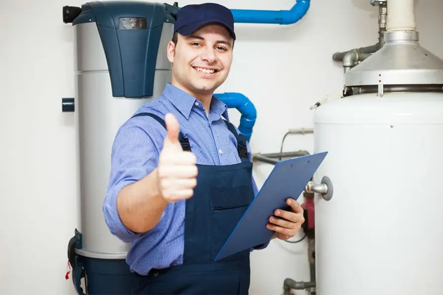 Young plumber in blue shirt and cap holding clipboard giving thumbs up gesture in front of water heater and pipes.