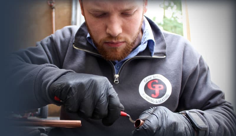 Skilled tradesman wearing black gloves and a gray G1 jacket performs precise welding on copper piping indoors.