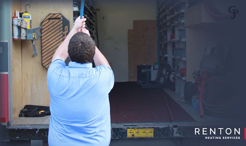 Technician in blue shirt reaching up to install equipment in residential garage with toolbox visible.
