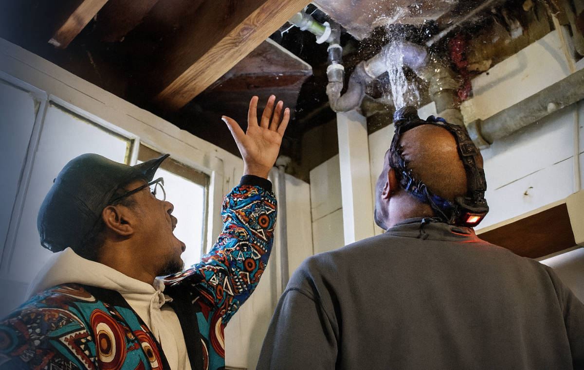 Gene Johnson plumber and homeowner examining water leak from ceiling pipe.